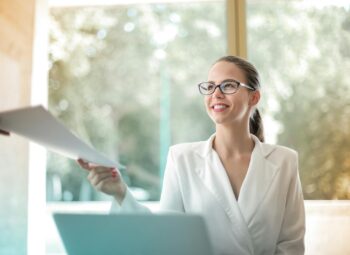 Low angle of successful female executive manager in classy style sitting at table with laptop in contemporary workplace and passing documents to colleague